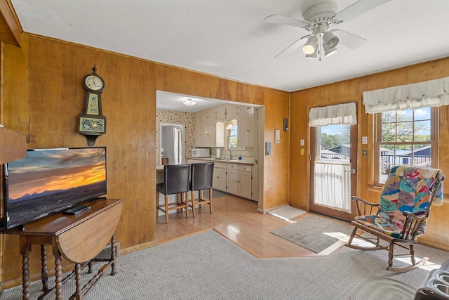 living area featuring light wood-type flooring, a ceiling fan, healthy amount of natural light, wood walls, and baseboards