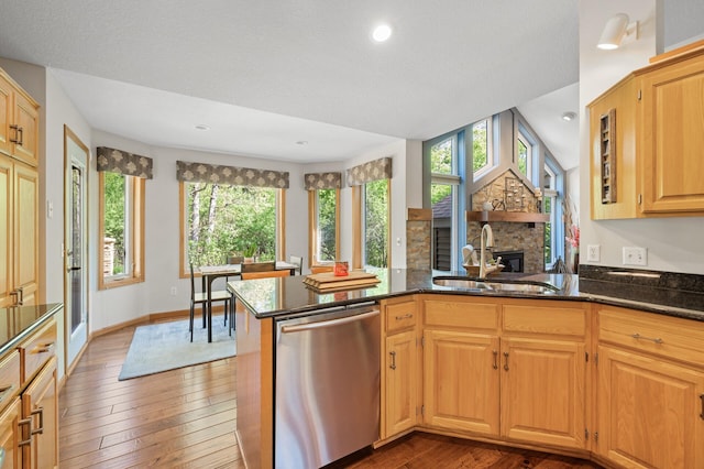 kitchen featuring dishwasher, a sink, wood-type flooring, dark stone counters, and a stone fireplace