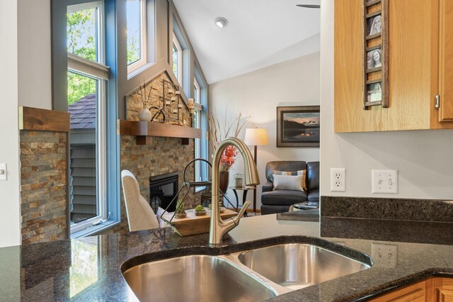 kitchen featuring a sink, a glass covered fireplace, dark stone counters, and vaulted ceiling