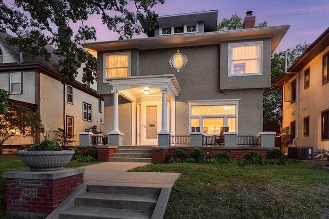 traditional style home featuring a chimney, stucco siding, a porch, a lawn, and cooling unit