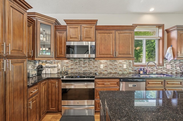 kitchen featuring appliances with stainless steel finishes, a sink, brown cabinetry, dark stone counters, and backsplash