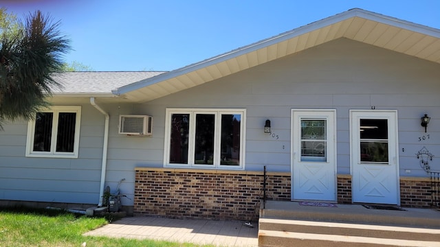 doorway to property featuring brick siding, a wall mounted AC, and roof with shingles