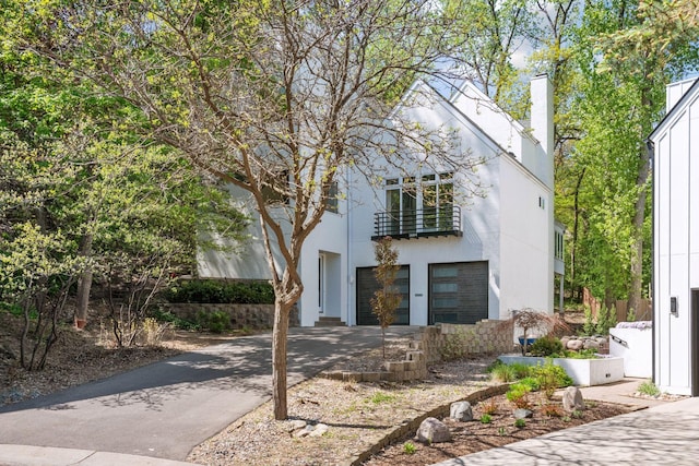 art deco inspired home featuring driveway, an attached garage, a chimney, a balcony, and stucco siding