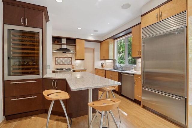 kitchen with stainless steel appliances, wall chimney exhaust hood, light wood-type flooring, backsplash, and recessed lighting