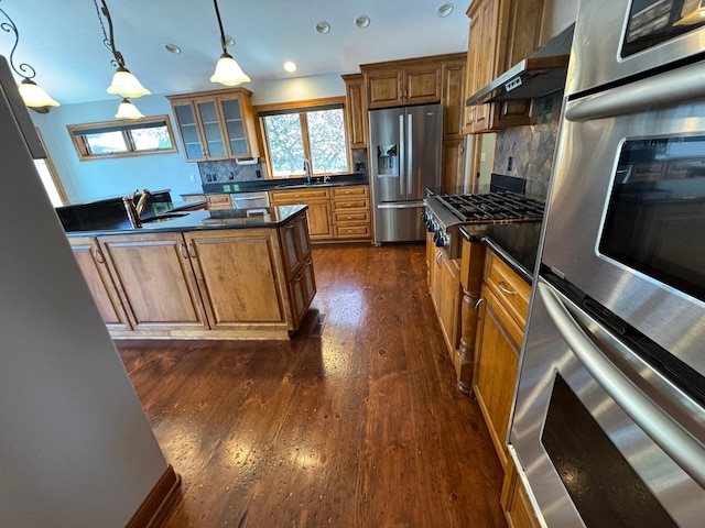 kitchen with stainless steel appliances, a sink, dark countertops, exhaust hood, and brown cabinetry