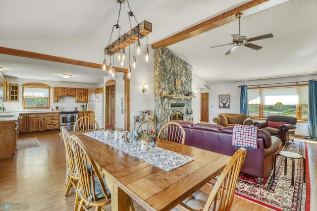 dining space featuring a ceiling fan, plenty of natural light, beam ceiling, light wood-type flooring, and a stone fireplace