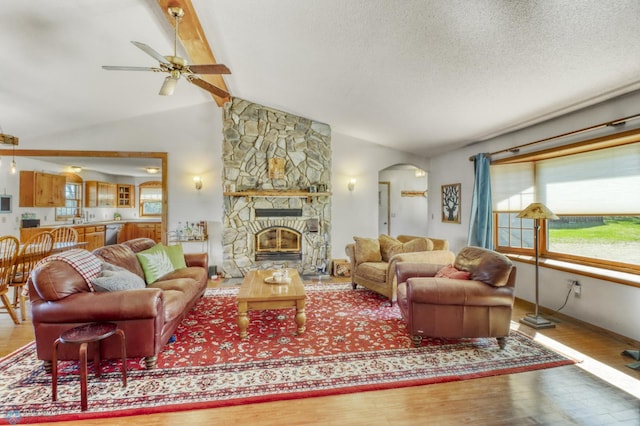 living area with arched walkways, wood finished floors, ceiling fan, a stone fireplace, and a textured ceiling