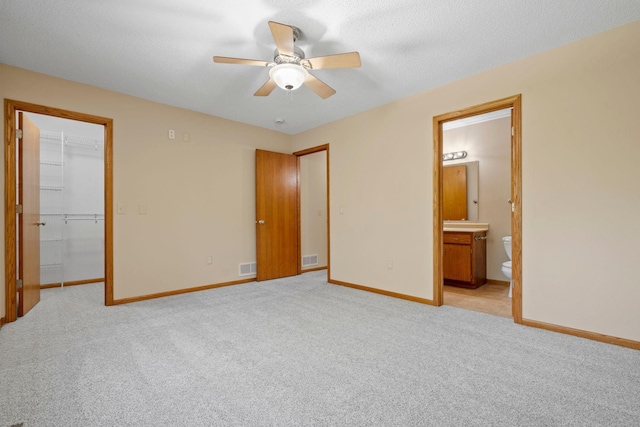 unfurnished bedroom with baseboards, a spacious closet, light colored carpet, ensuite bathroom, and a ceiling fan