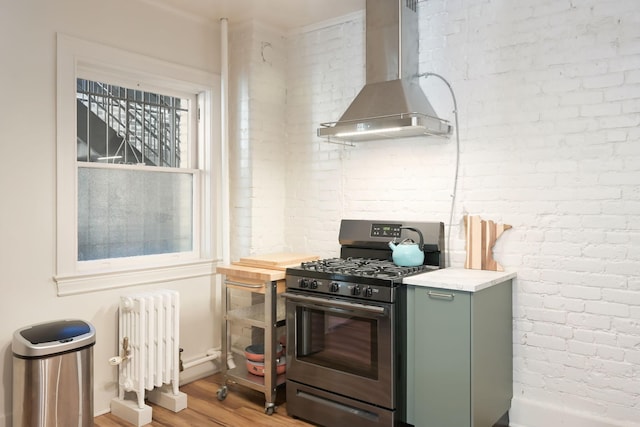 kitchen featuring stainless steel gas range oven, radiator, wall chimney exhaust hood, light wood finished floors, and green cabinetry