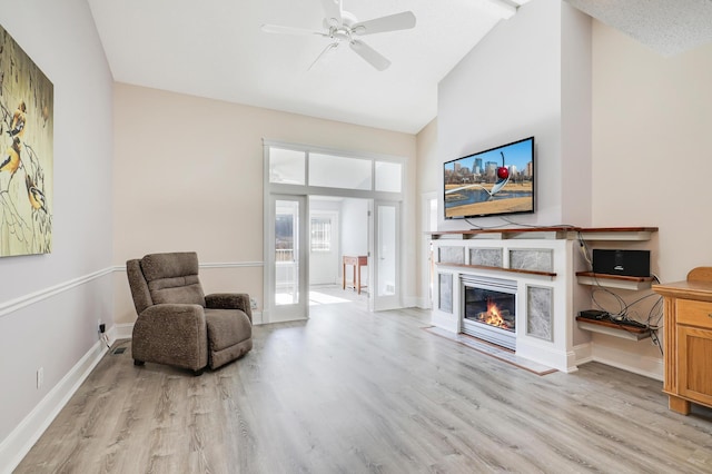 sitting room featuring light wood-style floors, high vaulted ceiling, a glass covered fireplace, ceiling fan, and baseboards
