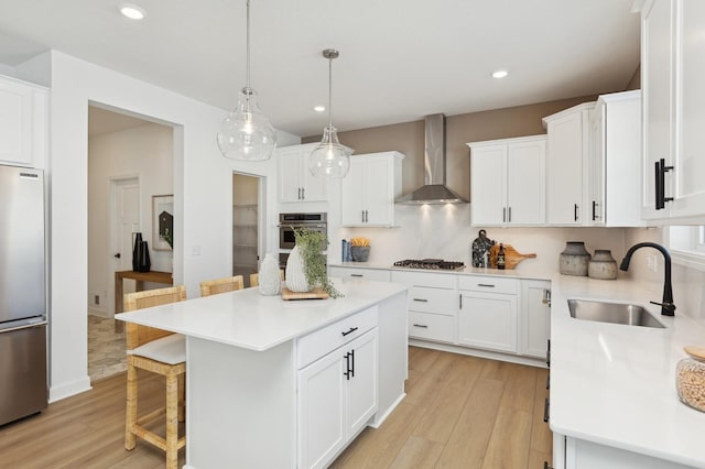 kitchen featuring wall chimney exhaust hood, stainless steel appliances, a sink, light wood finished floors, and a kitchen bar