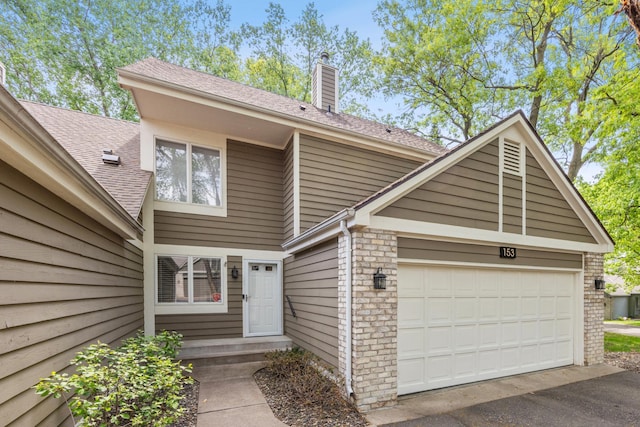 view of exterior entry with a chimney, an attached garage, driveway, a shingled roof, and brick siding