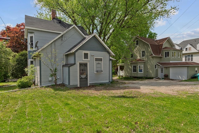 rear view of property featuring driveway, a chimney, a garage, and a lawn