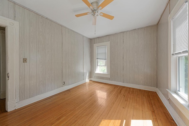 empty room with light wood finished floors, plenty of natural light, ceiling fan, wood walls, and baseboards
