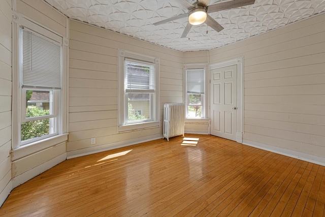 unfurnished room featuring radiator, plenty of natural light, ceiling fan, light wood-type flooring, and wood walls