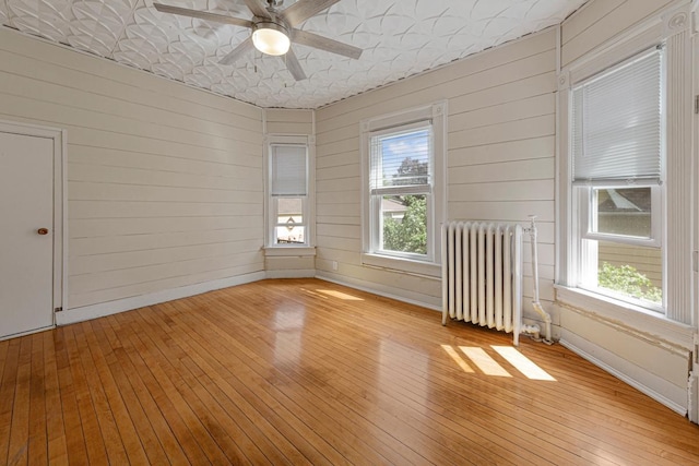 empty room with radiator heating unit, light wood-style flooring, wooden walls, a ceiling fan, and baseboards