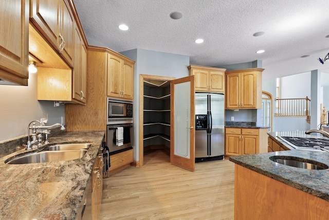 kitchen featuring appliances with stainless steel finishes, a sink, dark stone counters, light wood-style floors, and a textured ceiling
