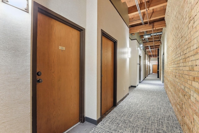corridor featuring brick wall, wood ceiling with exposed beams, light colored carpet, and baseboards