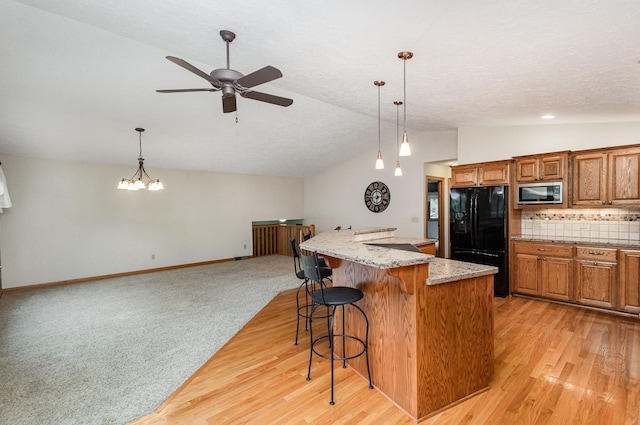 kitchen featuring freestanding refrigerator, vaulted ceiling, stainless steel microwave, brown cabinetry, and a kitchen bar