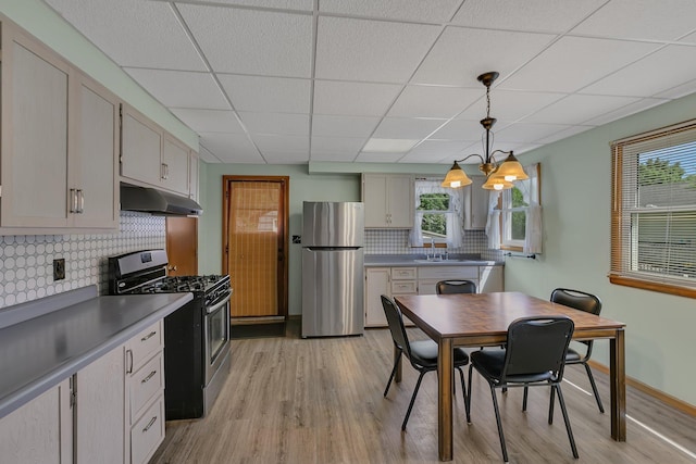kitchen with appliances with stainless steel finishes, under cabinet range hood, light wood finished floors, decorative backsplash, and a drop ceiling