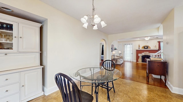 dining space featuring arched walkways, a chandelier, baseboards, a warm lit fireplace, and a ceiling fan