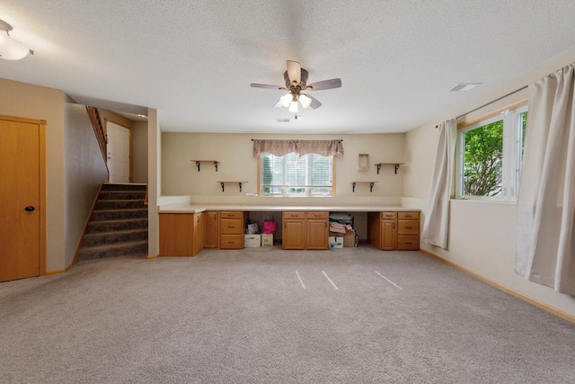 unfurnished office featuring healthy amount of natural light, built in desk, ceiling fan, a textured ceiling, and light colored carpet
