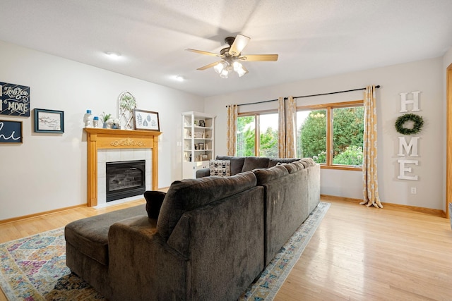 living room with a tiled fireplace, a ceiling fan, light wood-style flooring, and baseboards