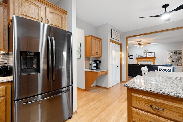 kitchen with stainless steel refrigerator with ice dispenser, a ceiling fan, a fireplace, light wood-type flooring, and light stone counters