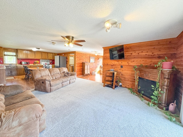 living area featuring wooden walls, a textured ceiling, ceiling fan, a glass covered fireplace, and light colored carpet