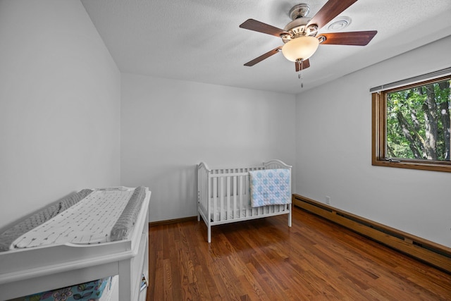 bedroom with baseboard heating, wood finished floors, a nursery area, ceiling fan, and a textured ceiling