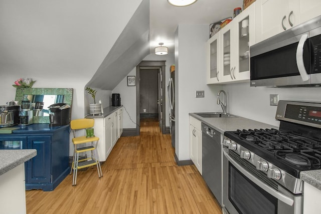kitchen with appliances with stainless steel finishes, a sink, white cabinetry, light wood finished floors, and glass insert cabinets