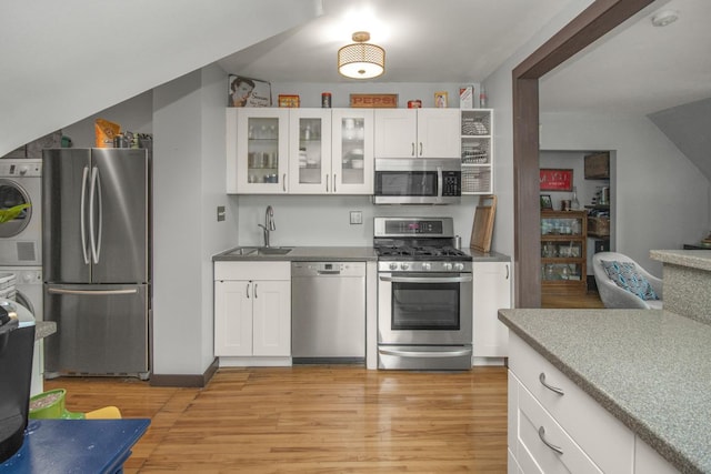 kitchen with appliances with stainless steel finishes, a sink, stacked washer / dryer, white cabinets, and light wood-type flooring