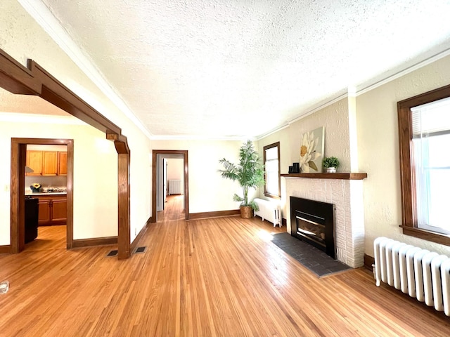unfurnished living room with radiator heating unit, a textured ceiling, light wood finished floors, crown molding, and a brick fireplace