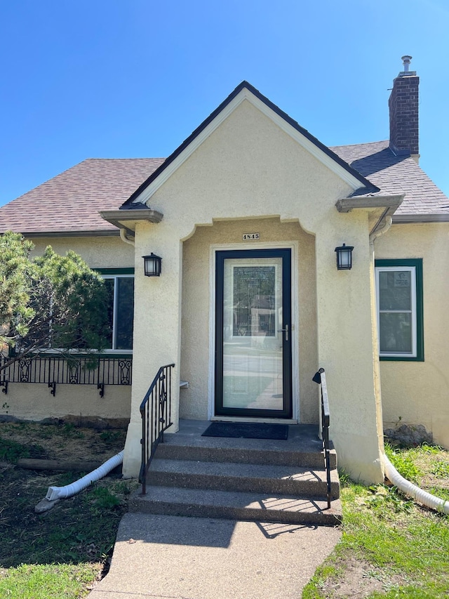 view of exterior entry with stucco siding, a chimney, and a shingled roof