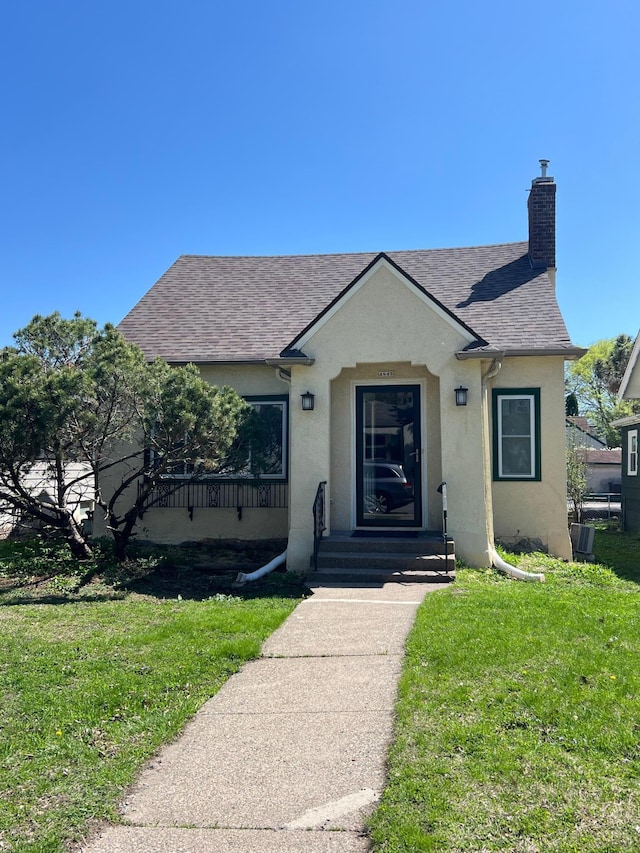 bungalow-style home with stucco siding, a front lawn, a chimney, and a shingled roof