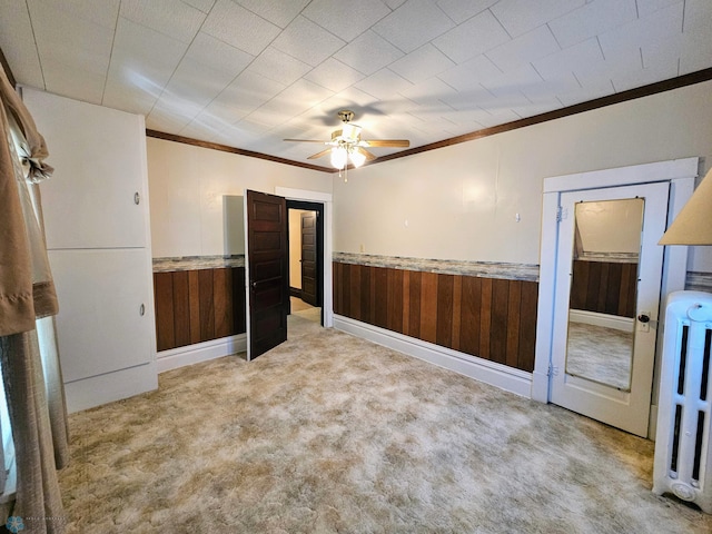 unfurnished bedroom with crown molding, wood walls, light carpet, wainscoting, and ceiling fan