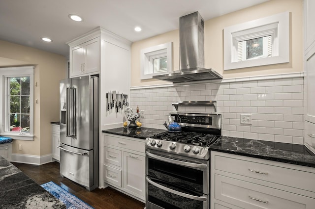 kitchen with stainless steel appliances, wall chimney exhaust hood, dark stone countertops, dark wood-style floors, and backsplash