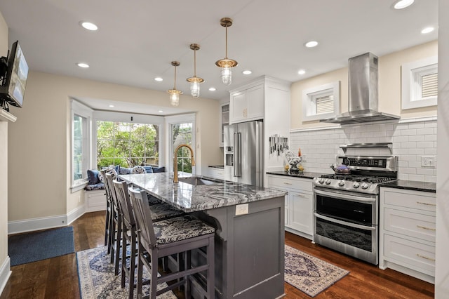 kitchen with wall chimney exhaust hood, stainless steel appliances, a sink, dark stone counters, and a kitchen breakfast bar