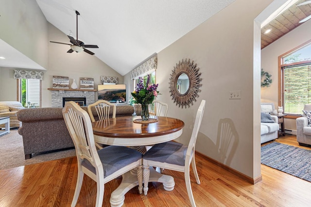 dining area featuring a ceiling fan, vaulted ceiling, a brick fireplace, light wood-style flooring, and baseboards