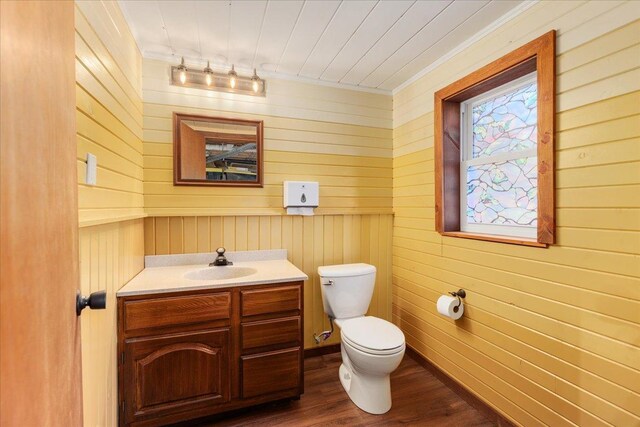 half bathroom with toilet, wood finished floors, vanity, wood walls, and ornamental molding