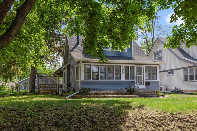 view of front facade with roof with shingles, a front yard, a wooden deck, and a sunroom