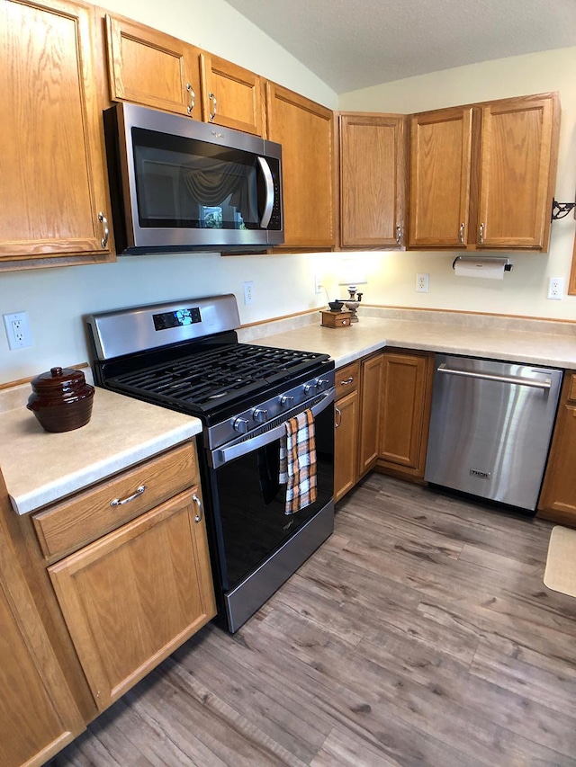 kitchen with appliances with stainless steel finishes, light countertops, wood finished floors, vaulted ceiling, and brown cabinets