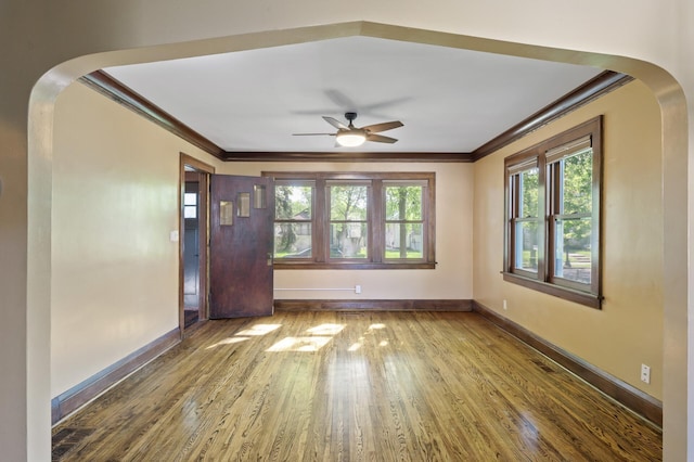 empty room with ornamental molding, a ceiling fan, arched walkways, wood finished floors, and baseboards