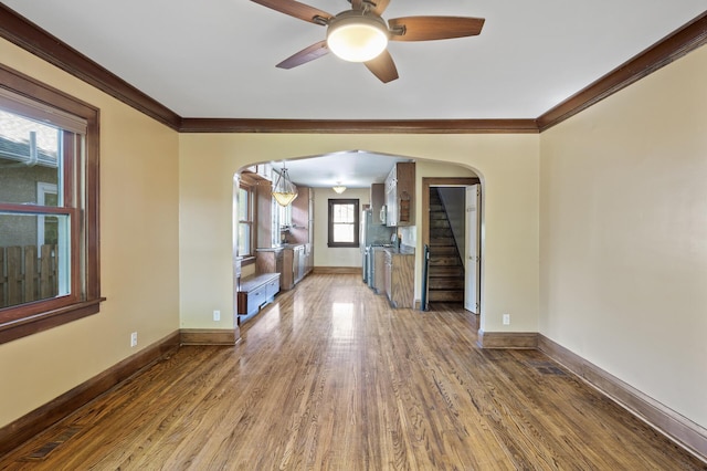 unfurnished living room featuring arched walkways, baseboards, wood finished floors, ornamental molding, and a ceiling fan