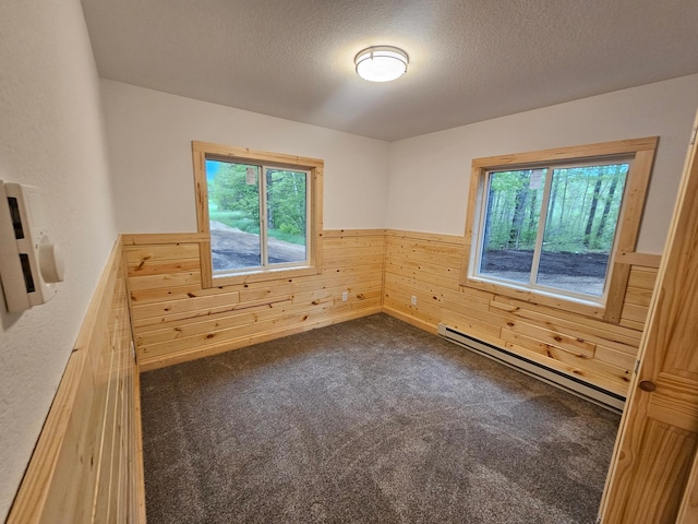 unfurnished room featuring a baseboard heating unit, wooden walls, healthy amount of natural light, wainscoting, and dark colored carpet