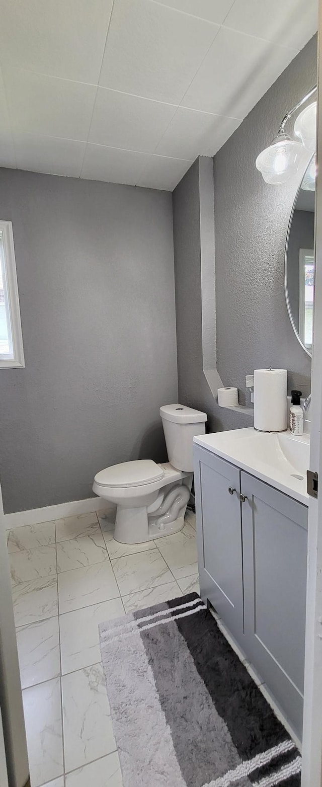 half bath with toilet, marble look tiles, vanity, a textured wall, and baseboards