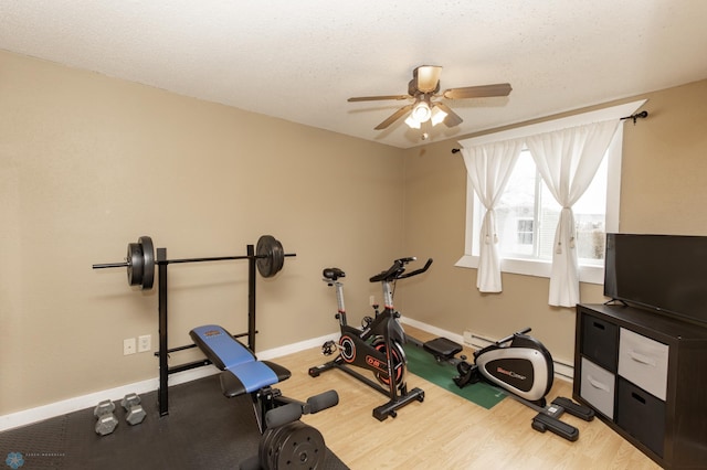 exercise area featuring a ceiling fan, wood finished floors, baseboards, a baseboard heating unit, and a textured ceiling