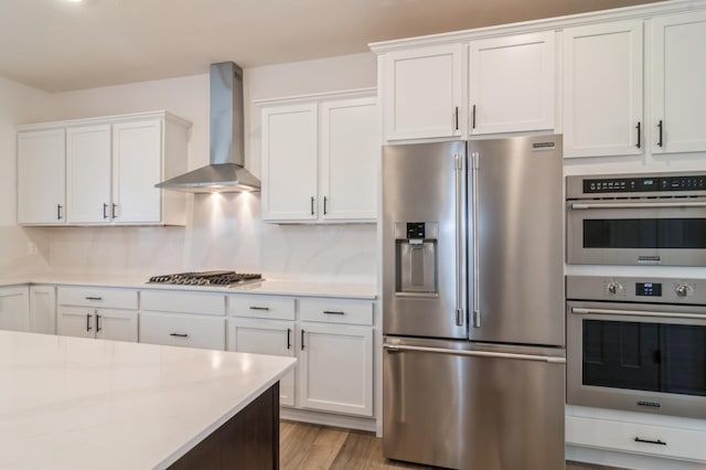 kitchen featuring stainless steel appliances, wall chimney exhaust hood, white cabinets, light wood finished floors, and tasteful backsplash
