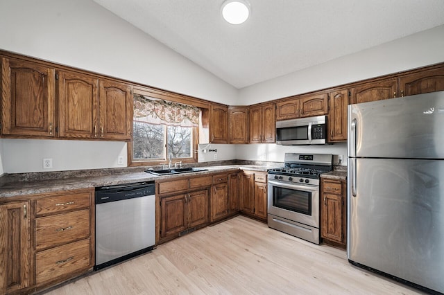 kitchen with appliances with stainless steel finishes, a sink, vaulted ceiling, light wood-type flooring, and brown cabinetry