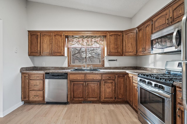 kitchen featuring appliances with stainless steel finishes, a sink, vaulted ceiling, light wood-style flooring, and dark countertops